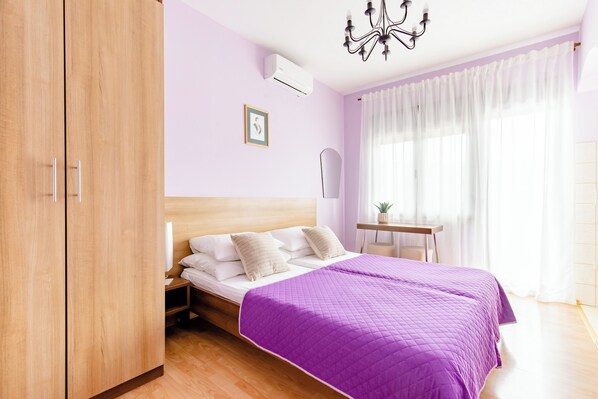 Un dormitorio con un armario de madera, una cama con colcha morada, una mesita de noche con lámpara y una ventana con cortinas traslúcidas.