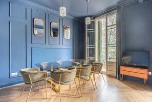 Ein Zimmer mit blauer Wand, einem Glastisch und vier Stühlen mit goldfarbenen Beinen. Ein Fernseher steht auf einem Holzständer und ein Fenster bietet Blick auf Bäume.