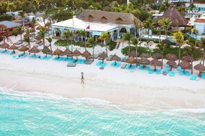 Un complejo turístico en la playa con un edificio de techo de paja, sillas de descanso y palmeras.