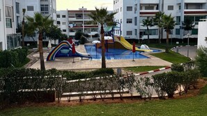 Un parque infantil con toboganes y una piscina, rodeado de palmeras y un edificio con balcones.
