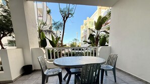 Un balcón con una mesa redonda y cuatro sillas, rodeado por una barandilla blanca, con edificios y árboles al fondo.