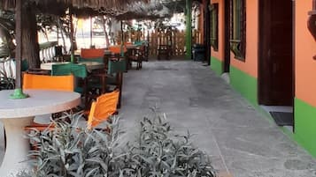 An outdoor seating area with tables and chairs, a thatched roof, and potted plants.
