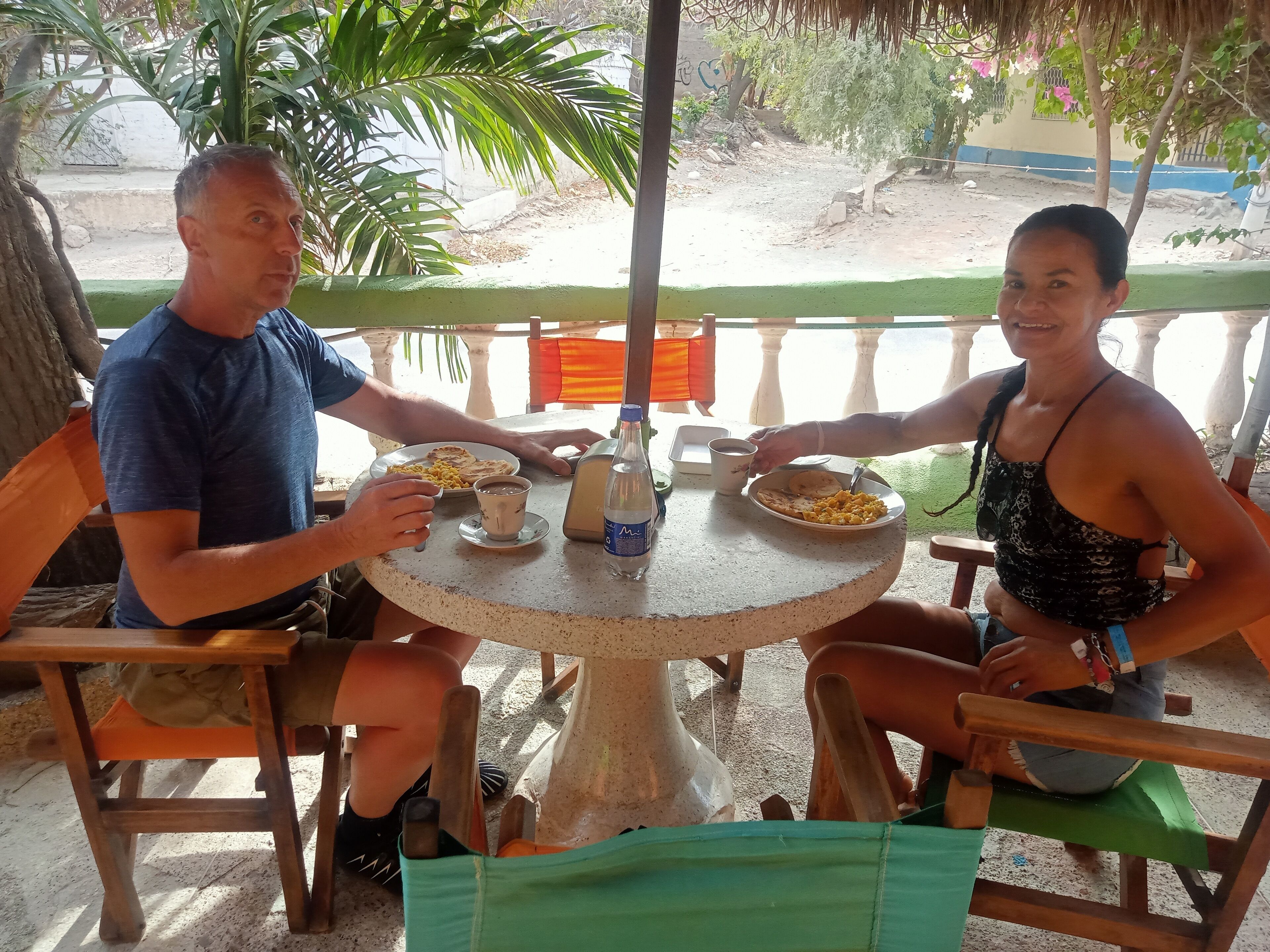 A man and a woman sitting at an outdoor table with plates of food, a bottle of water, and a cup.
