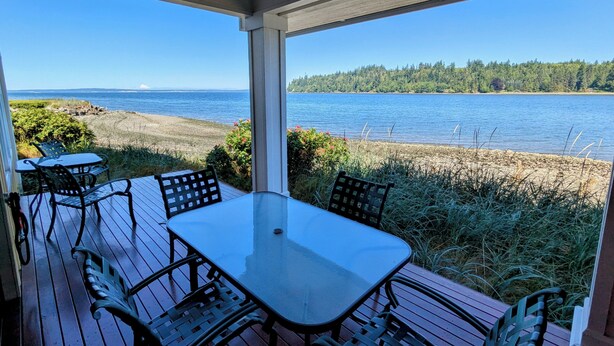A covered outdoor seating area with a table and chairs, overlooking a lake and forested shoreline. - Heron's Nest is a waterfront spectacular 3-bedroom townhome in Port Ludlow (Port Ludlow)
