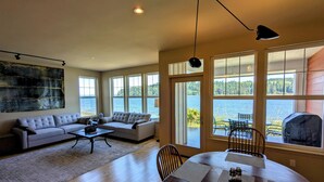 A spacious living room with a sofa, coffee table, and dining table. Large windows offer a view of a lake and outdoor seating. - Heron's Nest is a waterfront spectacular 3-bedroom townhome in Port Ludlow (Port Ludlow)