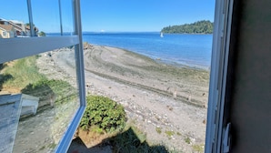 A view from a window overlooking a beach with a clear sky, a distant sailboat, and a forested island. - Heron's Nest is a waterfront spectacular 3-bedroom townhome in Port Ludlow (Port Ludlow)