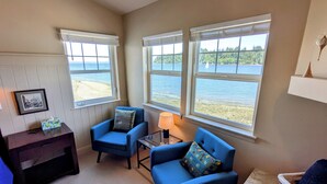 A room with two blue armchairs, a small table with a lamp, a framed picture on the wall, and large windows overlooking a beach. - Heron's Nest is a waterfront spectacular 3-bedroom townhome in Port Ludlow (Port Ludlow)