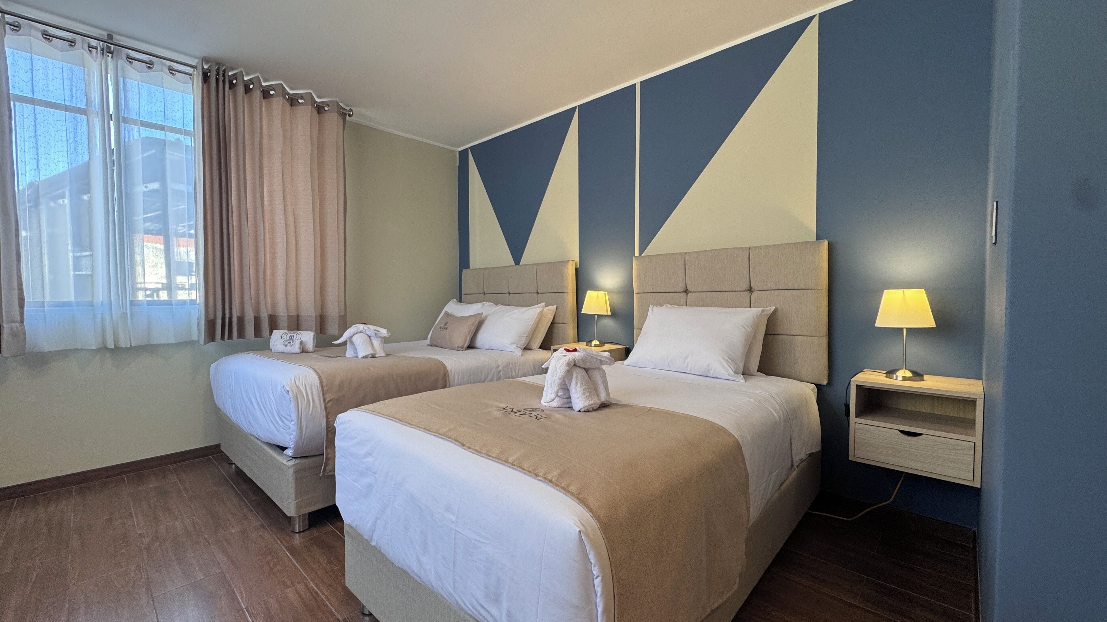 Una habitación de hotel con dos camas, una ventana con cortinas, una mesita de noche con lámpara y una pared con diseños geométricos.