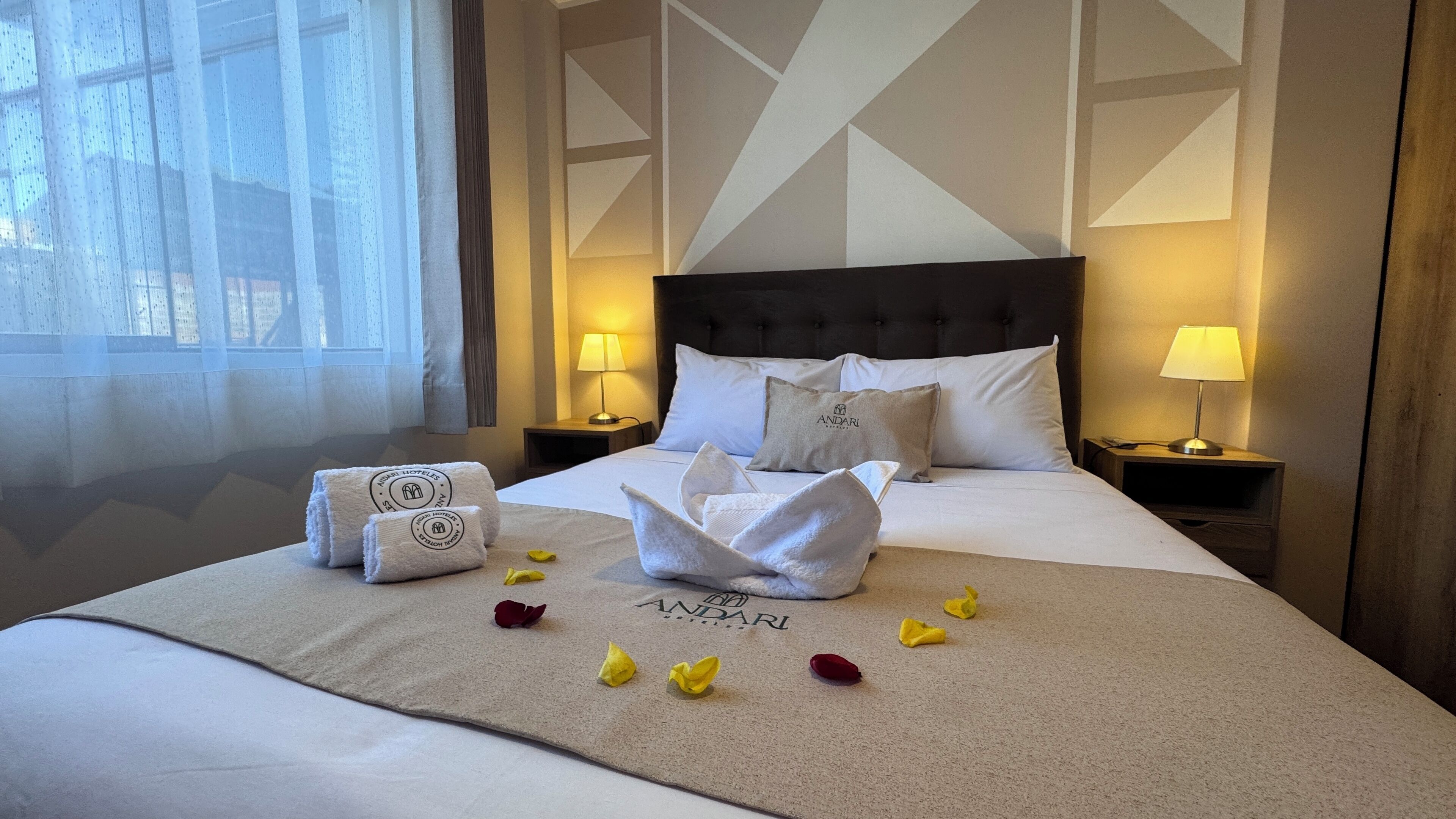 A hotel room with a neatly made bed, a headboard, two bedside tables with lamps, and a window with curtains.