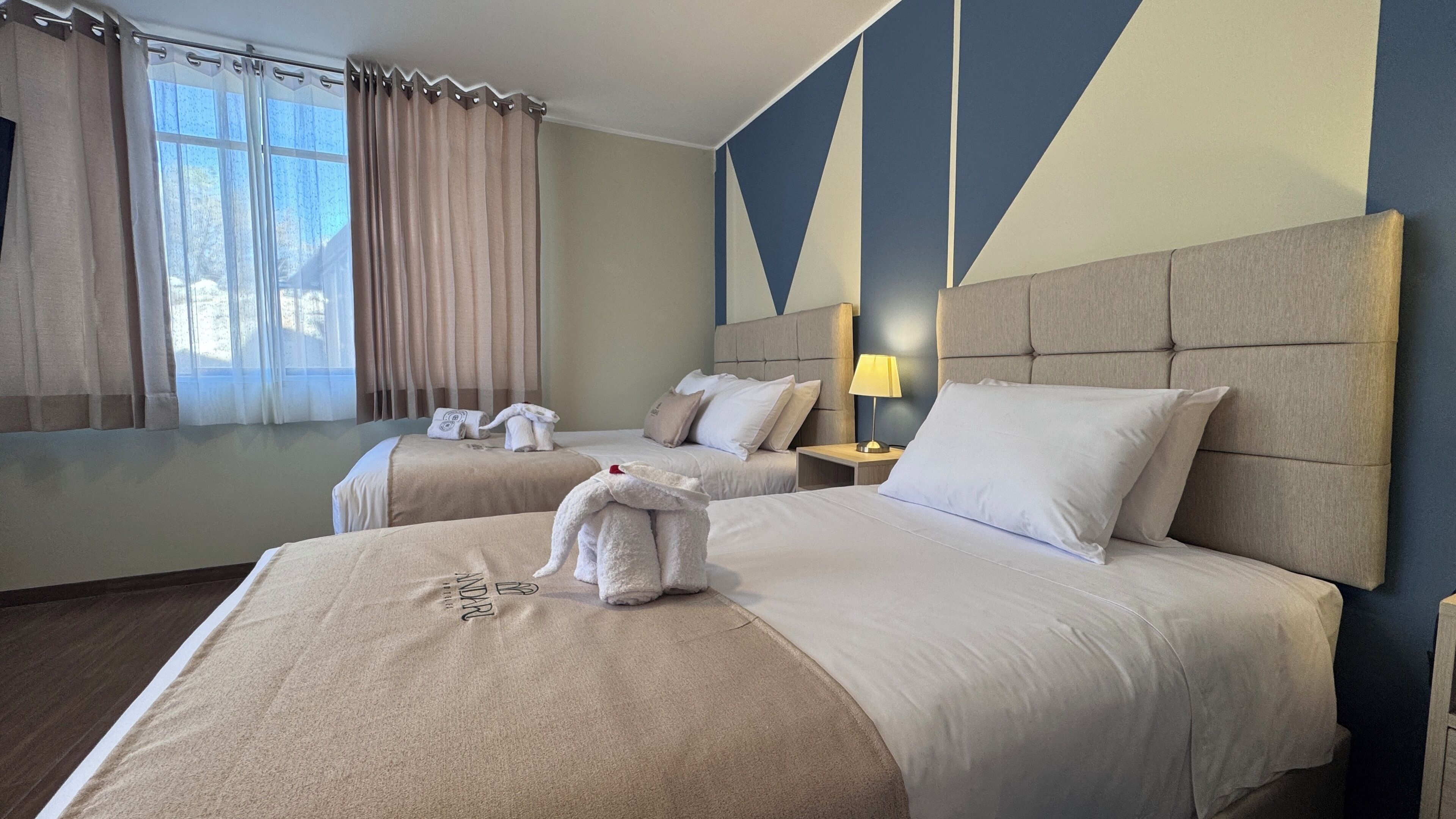 Ein Hotelzimmer mit zwei Betten, einem großen Fenster mit Vorhängen, einem Nachttisch mit Lampe und einer Wand mit geometrischen Mustern.