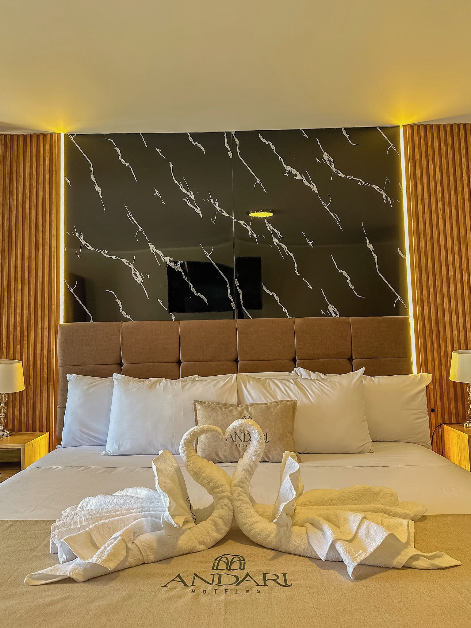 A hotel room with a bed, a headboard with a marble pattern, and a towel art display.