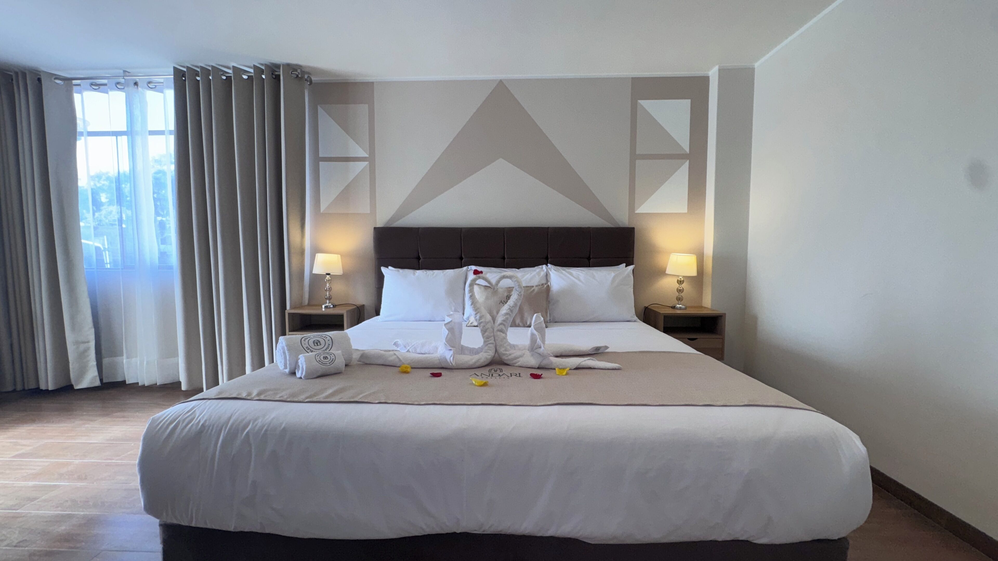 A hotel room with a large bed, two bedside tables with lamps, a window with curtains, and a geometric wall design.