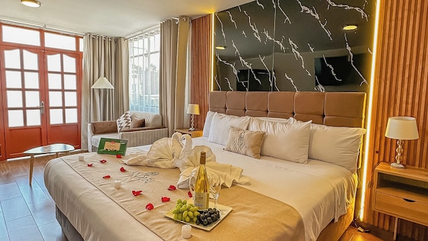A spacious hotel room with a large bed, a marble-patterned headboard, a bedside table with a lamp, and a view of the city through large windows. - ANDARI HOTELES (Arequipa)