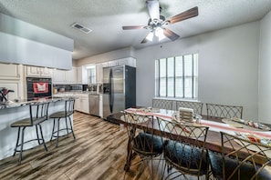 A modern kitchen with white cabinets, stainless steel appliances, a wooden dining table with chairs, and a ceiling fan.