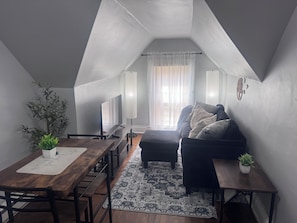 A cozy loft-style room with a dining area, a sofa, and a large window with sheer curtains.