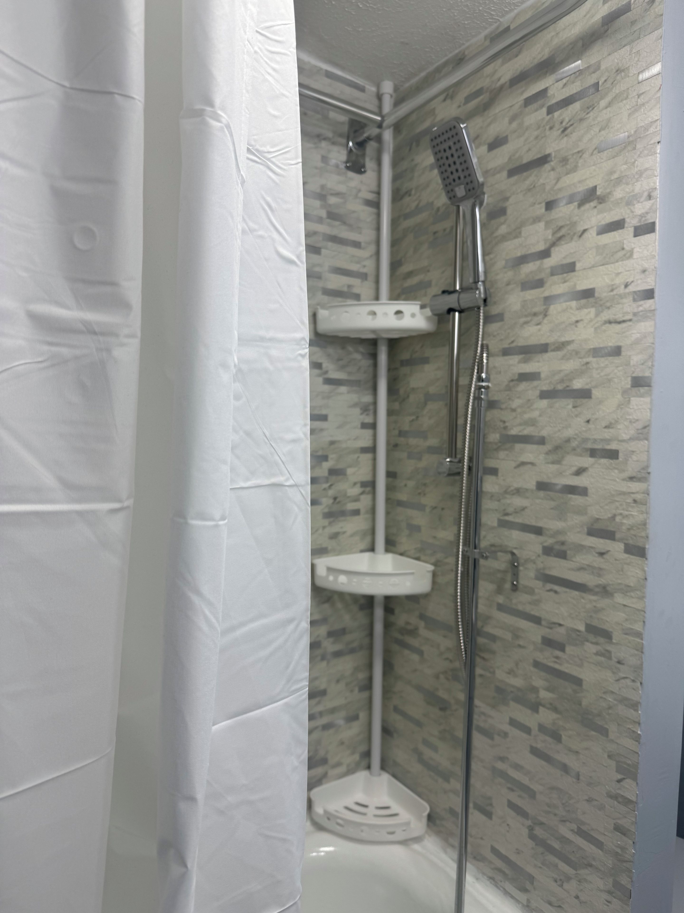 Un espace douche équipé d’un pommeau de douche à main, d’un robinet mural et d’une petite étagère blanche.