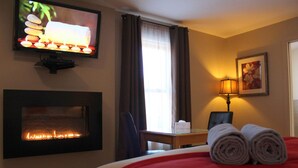 A hotel room with a fireplace, a bed with red bedding, a TV on the wall, a lamp, and a painting. - Auberge Aux Quatre Matins (Saint-Côme)