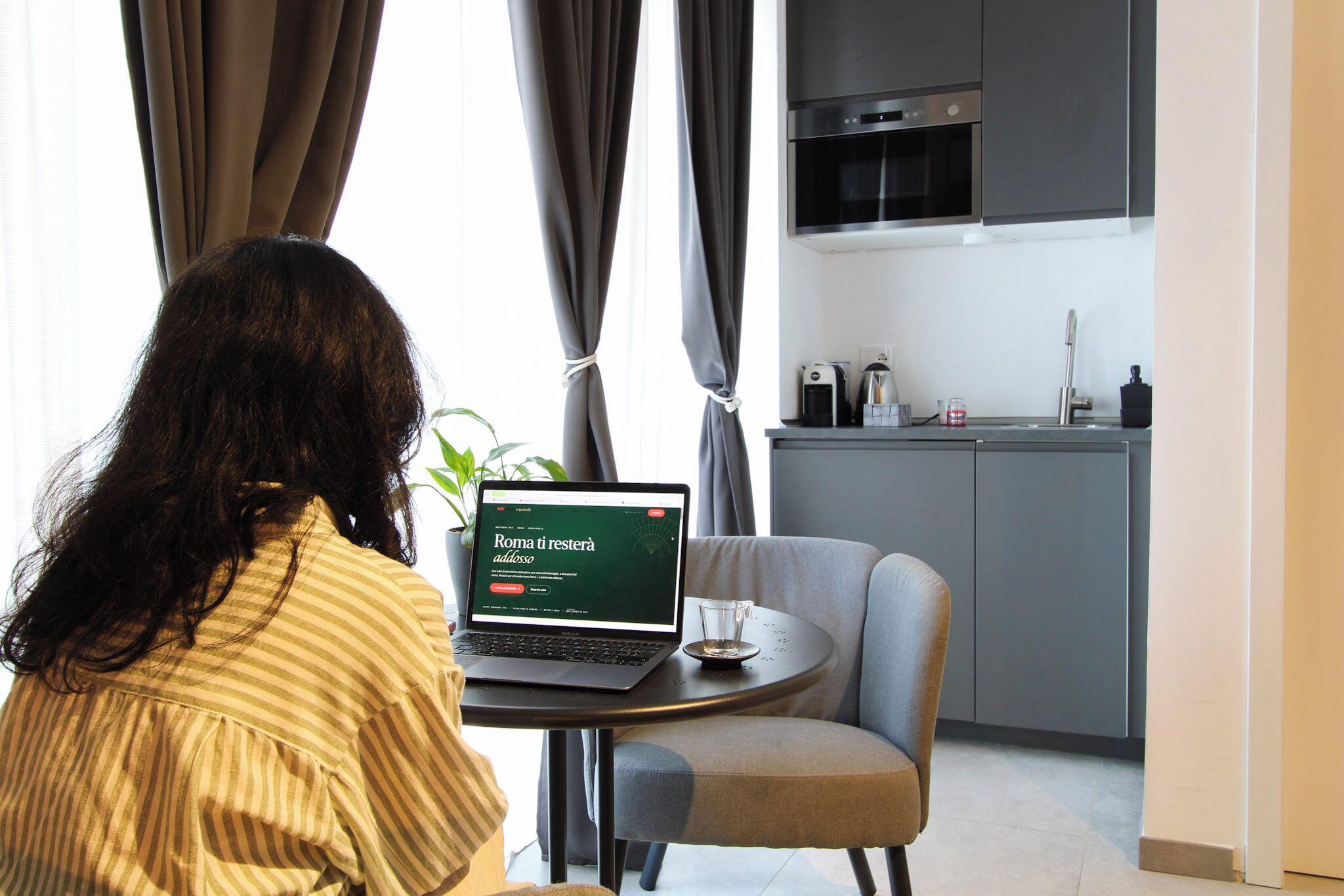 A person sitting at a table with a laptop, a glass of water, and a plant in a modern room with a kitchen area.