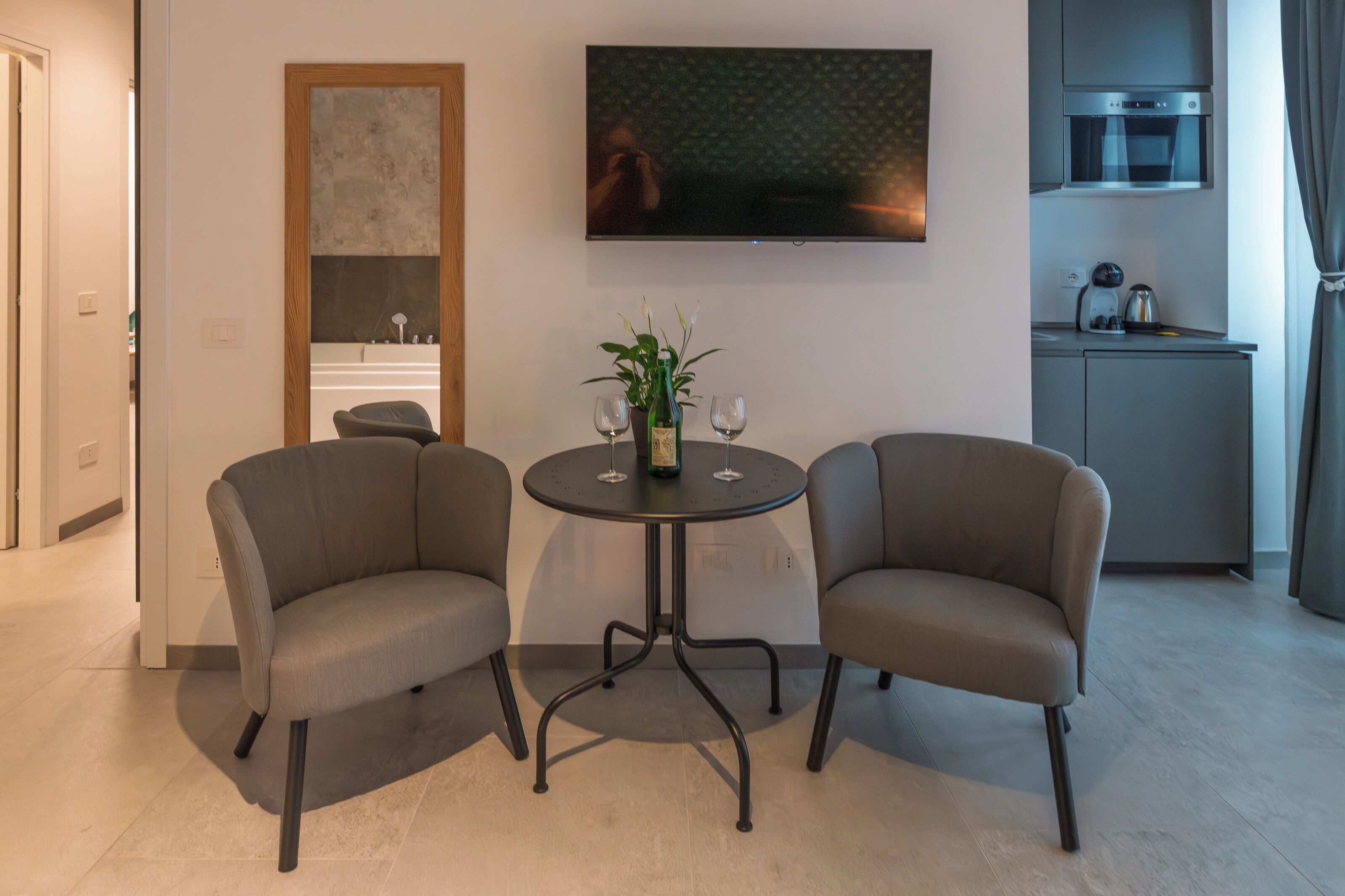 A modern living room with two sofas, a round table with a plant and wine glasses, a flat-screen TV, a kitchen area with a coffee machine, and a wooden-framed mirror.