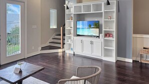 Una sala de estar moderna con un televisor de pantalla plana, estanterías blancas, una silla de mimbre y una escalera que lleva a un balcón.