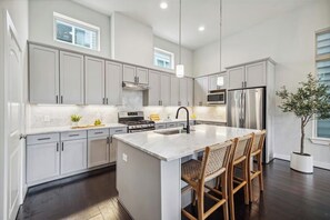 A modern kitchen with grey cabinets, a central island with bar stools, stainless steel appliances, and pendant lighting. - Modern Heights Home. 5 min to Downtown. Pet Friendly. (Houston)