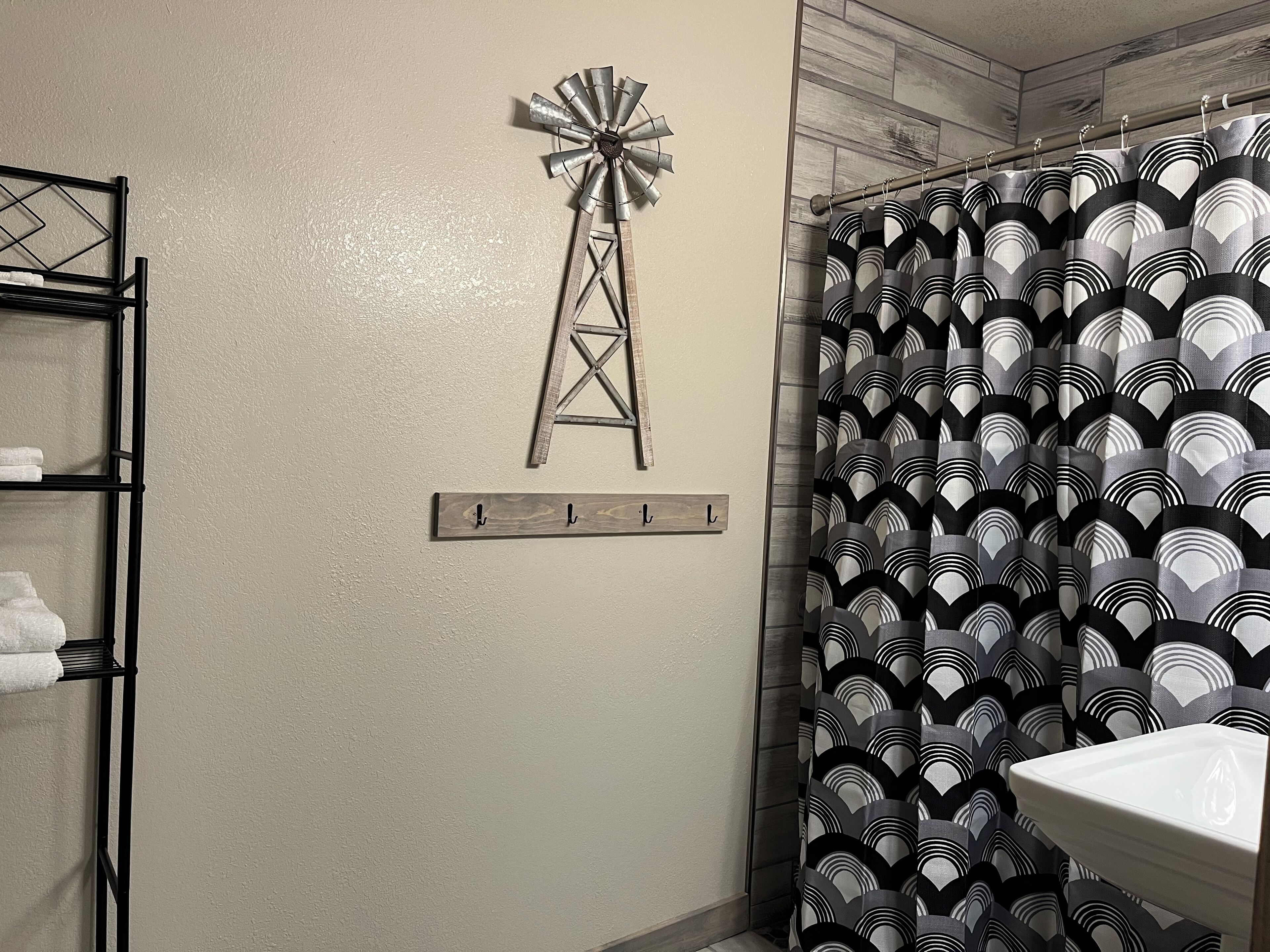 A bathroom with a shower curtain featuring a black and white geometric pattern, a wooden wall-mounted towel rack, and a metal windmill decoration.