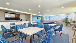 A modern lounge area with striped blue chairs, white tables, a pool table, a TV, and a large window offering a city view.