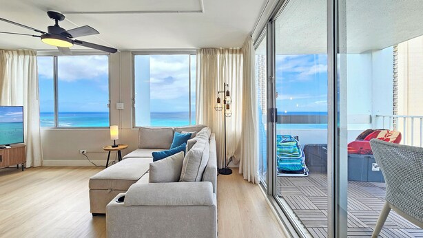 A spacious living room with a large sofa, a television, and a view of the ocean through large windows.