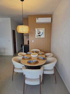 Uma sala de jantar com uma mesa redonda, seis cadeiras, um lustre pendente, um ar condicionado fixado na parede e uma obra de arte abstrata emoldurada.