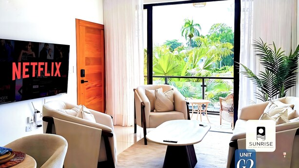 Un salon moderne doté d’un téléviseur à écran plat, de sièges confortables, d’un balcon offrant une vue sur la verdure et d’une plaque indiquant le numéro de l’appartement.