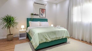 A modern bedroom with a teal bed, white bedding, a potted plant, and a bedside table with a lamp. - New Cairo Lush & Light (محافظة القاهرة‬)
