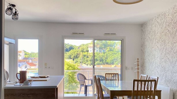 A modern kitchen with a wooden island, a dining area with wooden chairs, and a large sliding glass door leading to a balcony with a view of greenery and buildings. - Renovated two-bedroom flat (55m²), south-facing balcony  et  parking – town centre (ARCACHON)