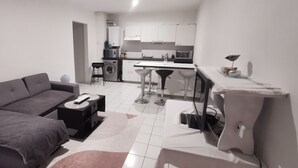 Un salon moderne avec un canapé gris, une petite table d’appoint noire et une table basse blanche. On aperçoit, au fond, une cuisine avec des meubles blancs et un four à micro-ondes.