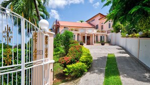 A large, two-story house with a red roof, surrounded by lush greenery and a white fence with decorative elements.