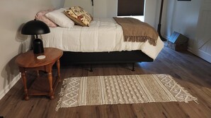 A bedroom with a bed, a bedside lamp, a wooden nightstand, a woven rug, a window with blinds, and a coat rack with a picture. - Brand new studio apartment with queen bed, queen pull out, WIFI and AC . (Wooster)