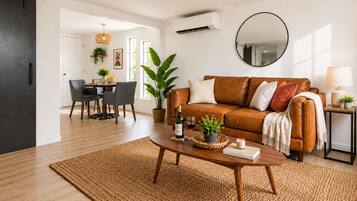 A modern living room with a brown leather sofa, a round mirror, a dining area with a table and chairs, and a large potted plant.