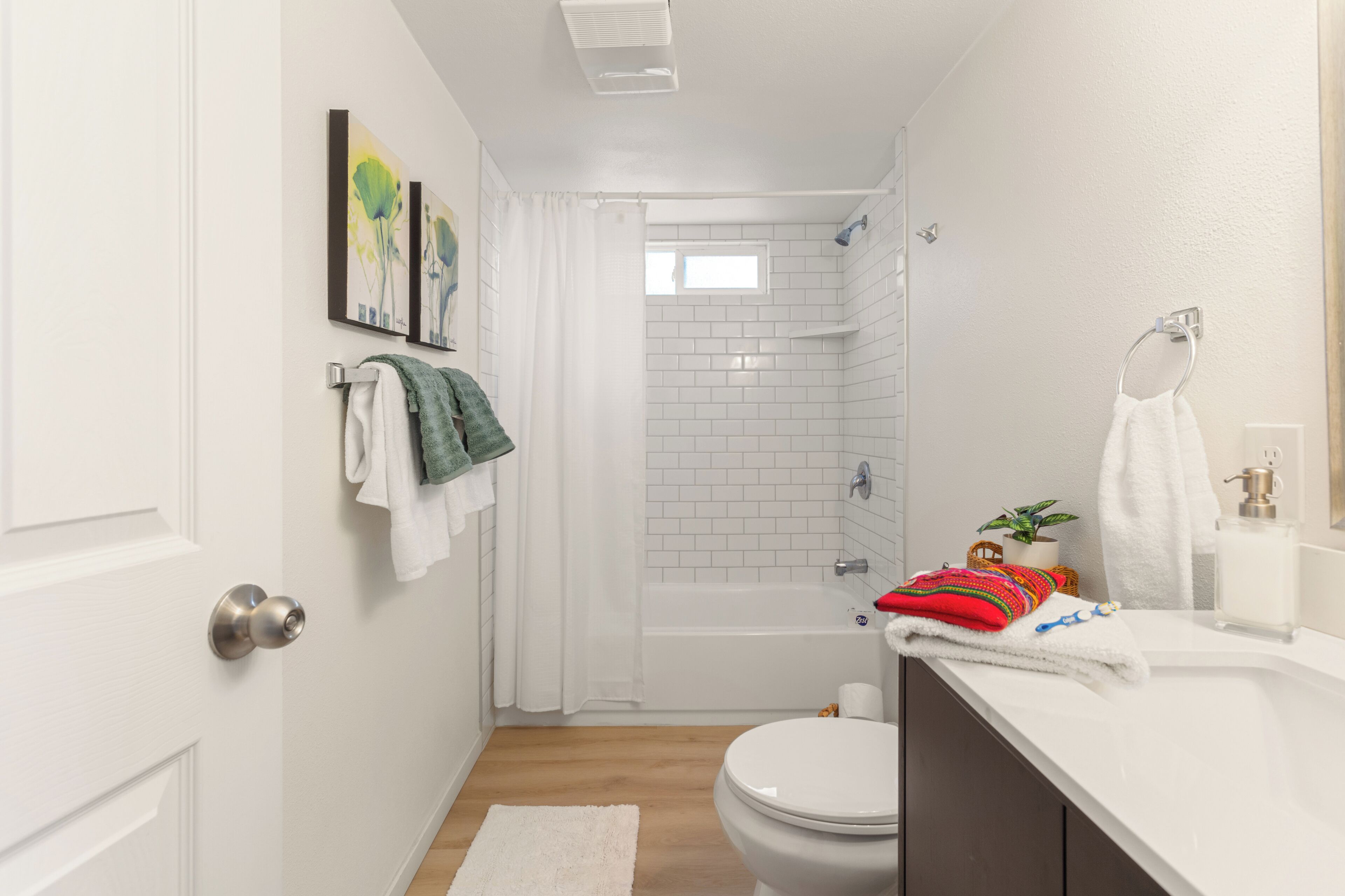 A modern bathroom with a white toilet, a shower, and a sink with a mirror. There are towels hanging on the wall and a small plant on the sink.
