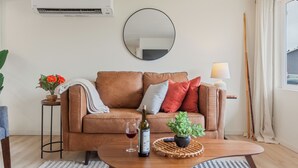 A living room with a brown leather sofa, a round mirror, a small table with a wine bottle and glass, and a potted plant.