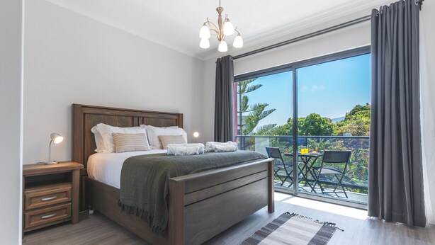 Un dormitorio con una cama grande, cabecero de madera, mesitas de noche y vistas a un balcón con mobiliario exterior.
