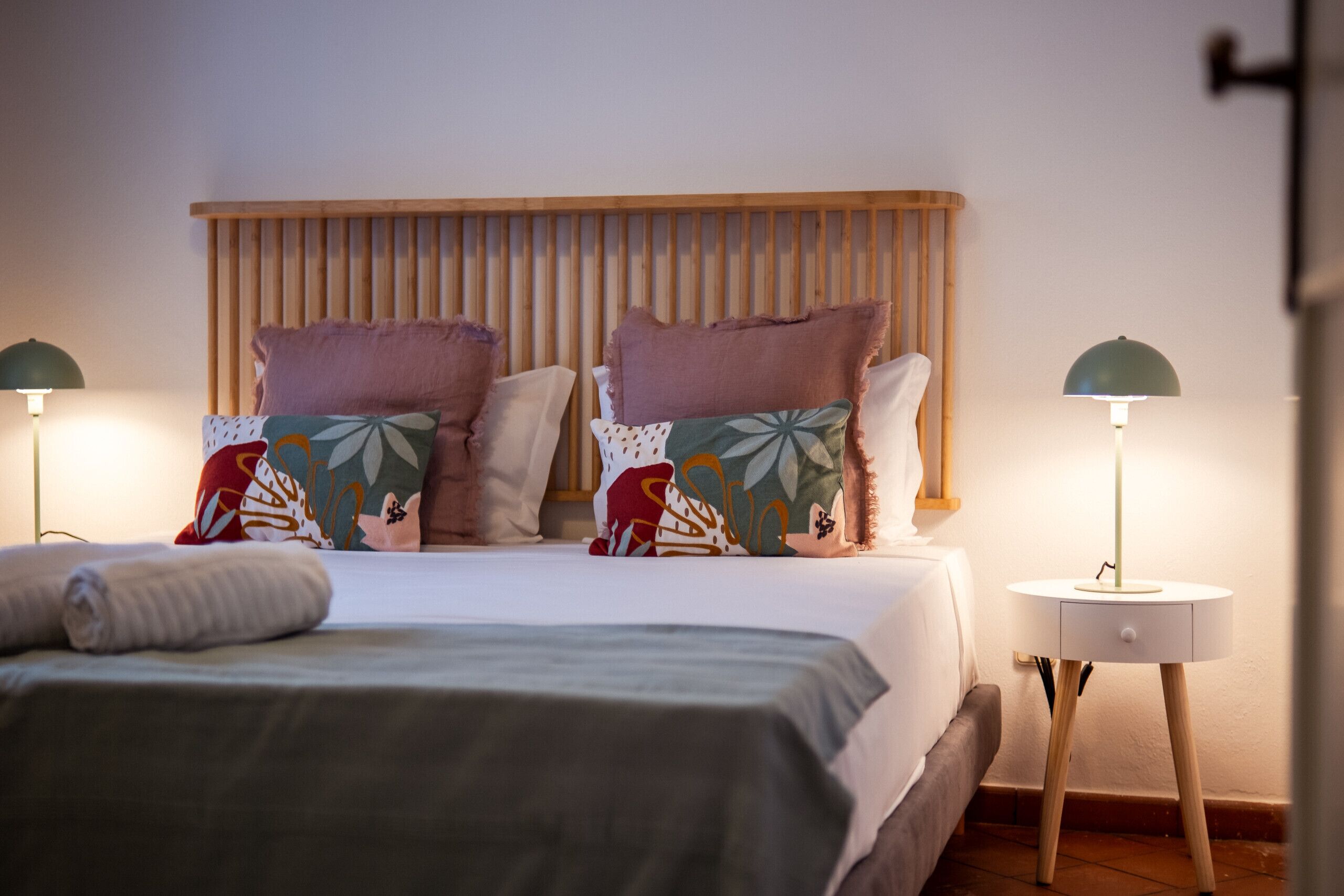 Un lit bien fait, avec une tête de lit en bois, deux tables de chevet avec des lampes et une variété d’oreillers à motifs.