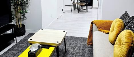 Una sala de estar moderna con una mesa de centro amarilla, un sofá con cojines color mostaza y un televisor. Se ve una cocina al fondo con armarios blancos y un microondas.