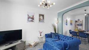 A modern living room with a large flat-screen TV, a blue sectional sofa, and a dining area with a table and chairs.
