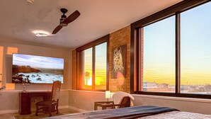A hotel room with a bed, a desk, a chair, a television, and large windows offering a city view.