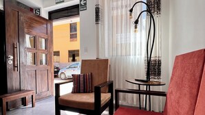 Un salon avec un canapé rouge, une chaise en bois, une petite table et un banc. On y trouve une porte avec une pancarte indiquant la sortie.