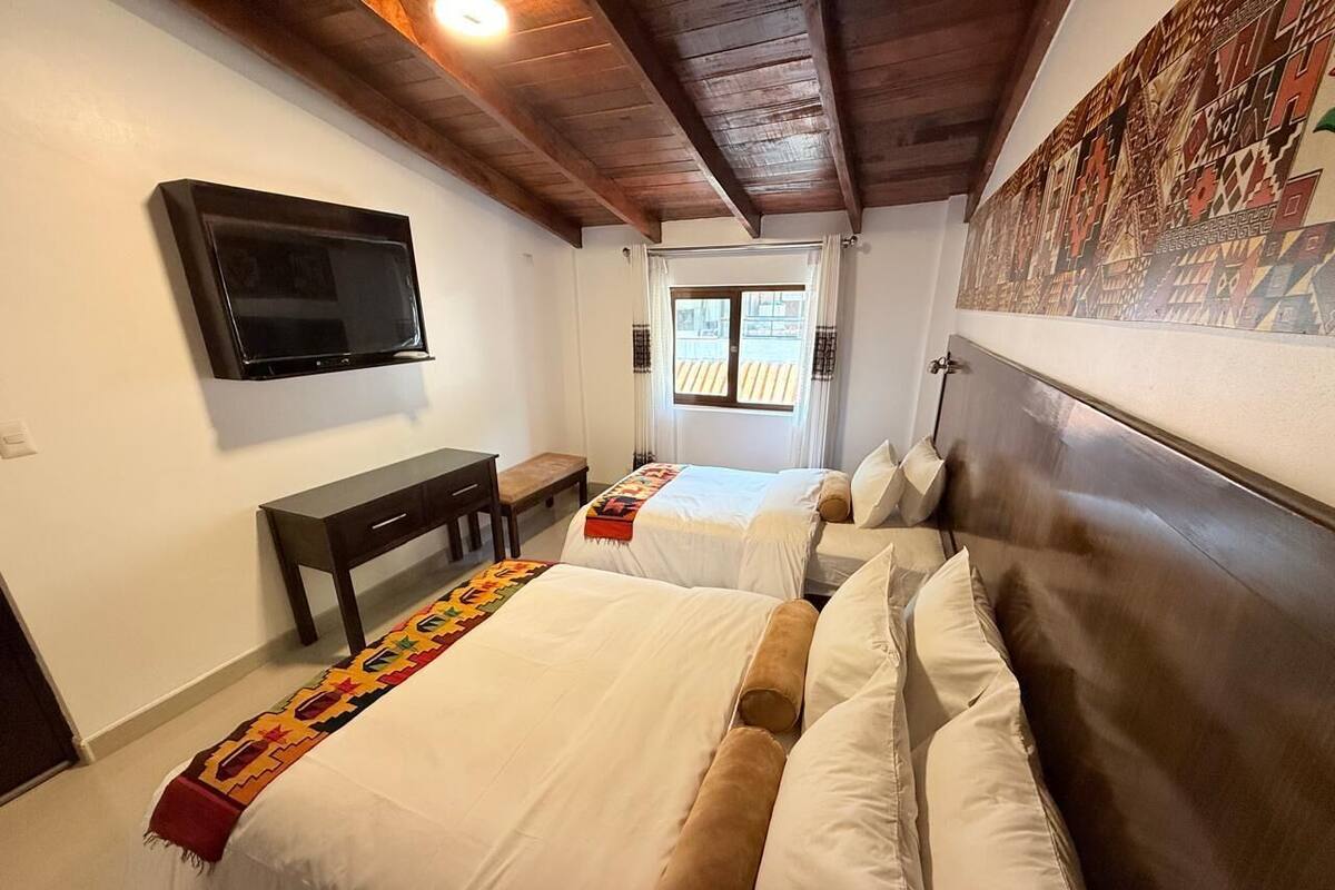 A hotel room with two beds, a TV mounted on the wall, a small desk, and a window with curtains.