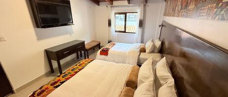 Habitación de hotel con dos camas, un televisor montado en la pared, un escritorio pequeño y una ventana con cortinas.