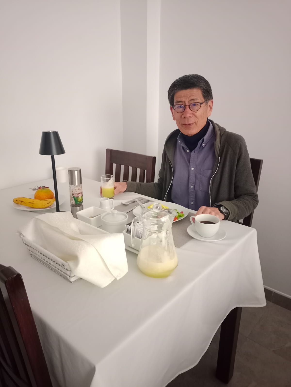 Ein Esstisch mit einer Karaffe, einem Glas Orangensaft, einer Banane, einer Kaffeetasse und einer weißen Tischdecke.