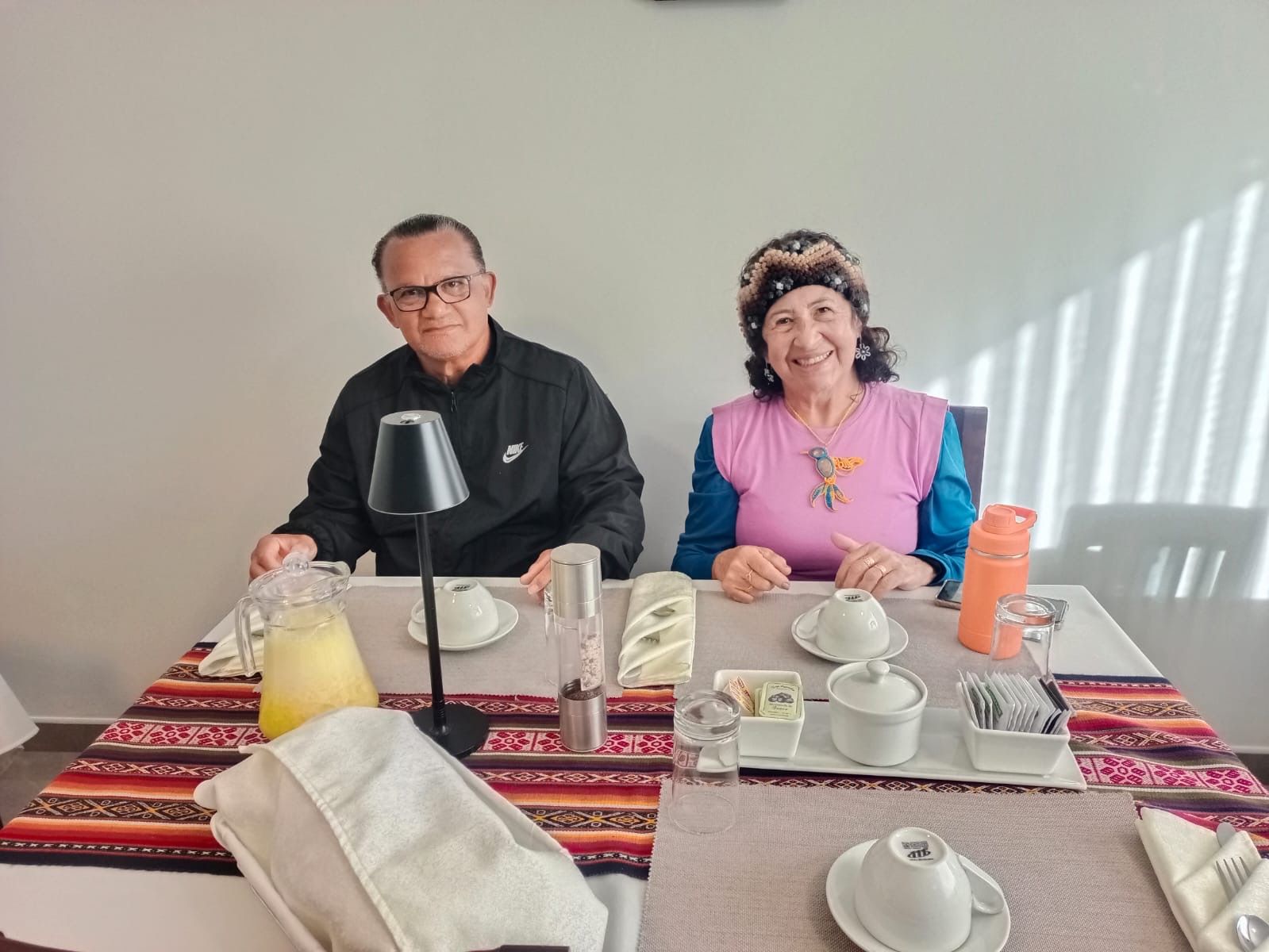 Dues persones assegudes a taula, amb un corredor de taula colorit, una làmpada i diferents elements de servei, com ara tasses, una teiera i una ampolla d'aigua.