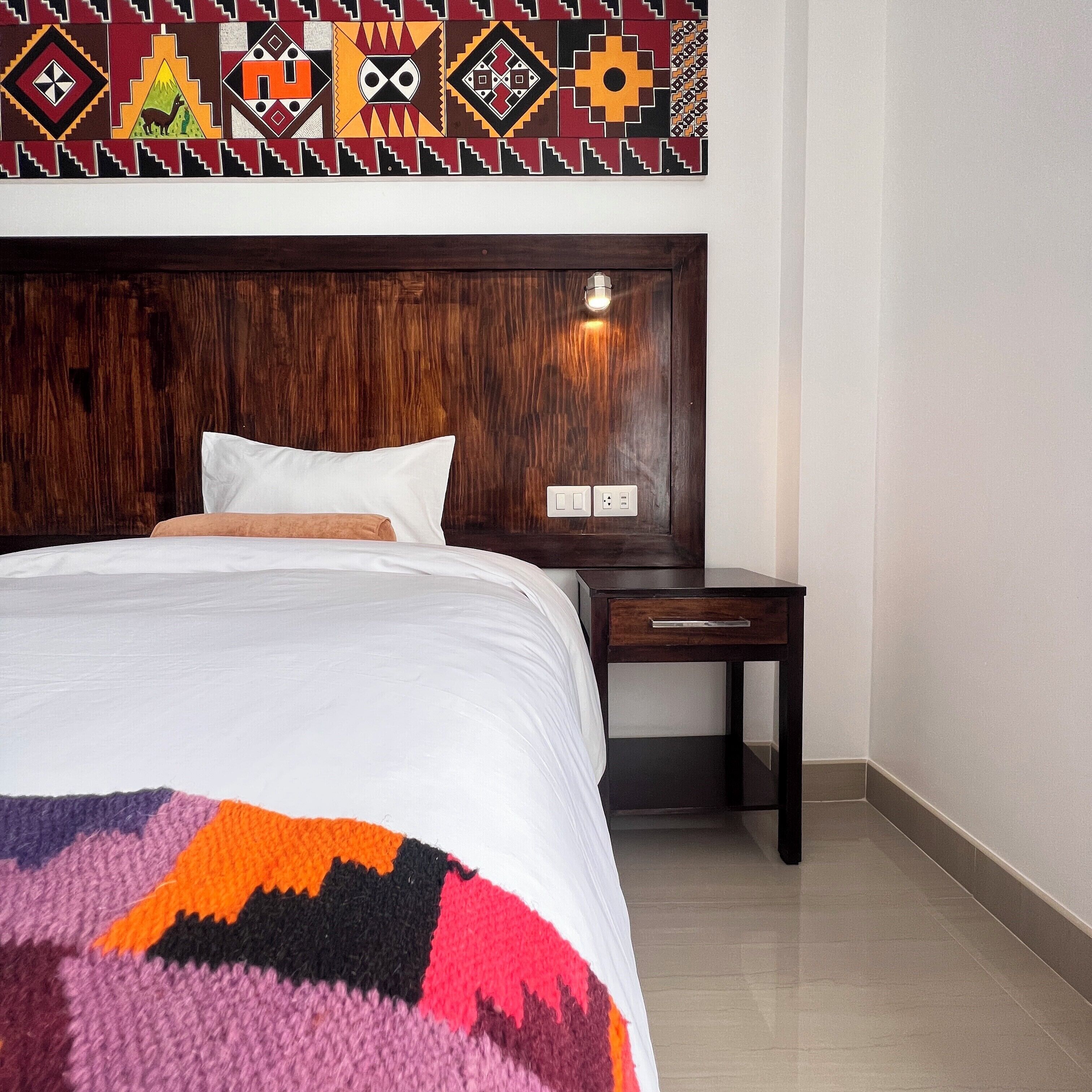 A hotel room with a wooden headboard, a white bedspread, a colorful blanket, a wooden nightstand, and a wall-mounted light fixture.