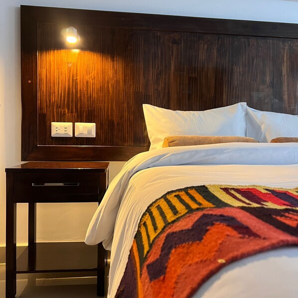 Une chambre d’hôtel avec une tête de lit en bois, une table de chevet et un lit recouvert de draps blancs et d’une couverture colorée.